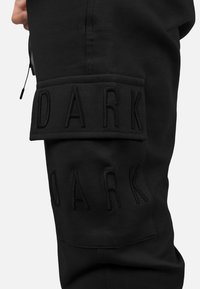 Los pantalones jogger negros presentan un diseño destacado en relieve con la palabra "DARK" en el muslo izquierdo, fabricado con tela texturizada y múltiples bolsillos utilitarios.