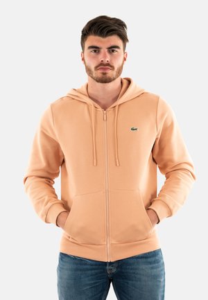 Perzikkleurige hoodie met rits en een capuchon met trekkoord, twee steekzakken aan de voorkant en een klein groen logo op de linkerborst. Zacht materiaal, casual pasvorm.