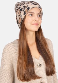 Bonnet en tricot beige avec un imprimé léopard noir et marron. Texture douce, bord roulé et coupe ajustée. Porté avec un pull chunky de couleur claire.