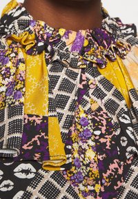 Blouse à motif floral offrant des couleurs jaunes, violettes et noires, avec un col volanté et un tissu texturé dans un design en patchwork.