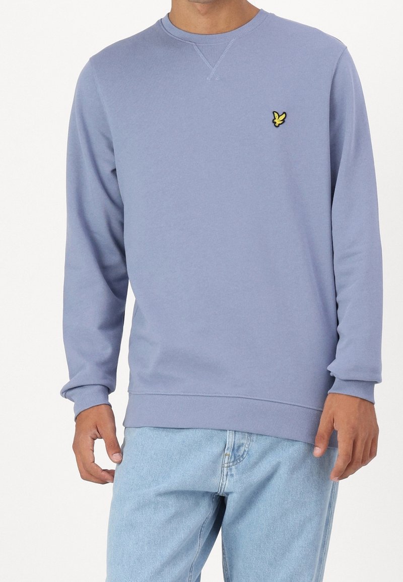 Ljusblå sweatshirt med rund halsringning, ribbade ärmslut och nederkant, med en liten gul broderad logotyp på vänster bröst. Avslappnad design.