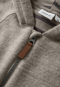 Sweat à capuche zippé beige avec une texture douce, présentant un tirette en cuir marron et un intérieur en tissu rayé.
