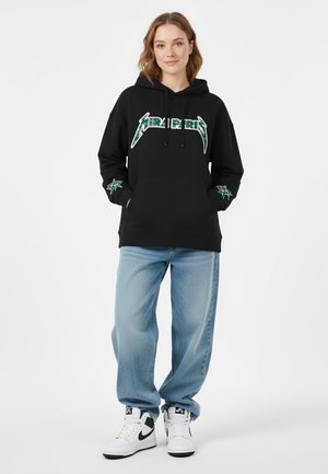 Haut à capuche noir avec texte graphique vert, comportant une poche kangourou et un cordon de serrage. Paire de jeans bleus clairs baggy et baskets blanches.