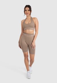 Bruine sportoutfit met een haltercrop top en high-waisted shorts, gemaakt van een gladde stof. Witte logo op de tailleband. Witte sneakers.