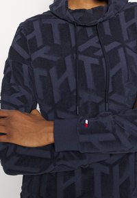 Marinblå hoodie med en strukturerad mönster, som har en huva med dragsko och ribbade ärmslut. Liten logotyp i röd, vit och blå på ärmen.