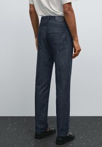 Una persona sta in piedi indossando pantaloni in denim scuro e scarpe nere, vista da dietro su uno sfondo neutro.