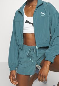 Turkis lynlåstrøje med elastiske manchetter, kombineret med en hvid sports-bh med et sort logo og matchende turkise shorts med snor.