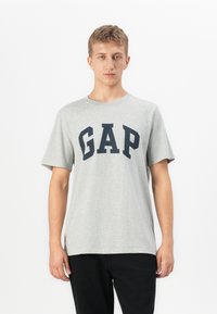 Ung mand iført en lysegrå T-shirt med korte ærmer, hvor "GAP" står skrevet i store marineblå bogstaver, parret med sorte bukser, stående mod en hvid baggrund.