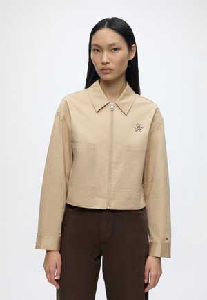 Frau mit glatten schwarzen Haaren trägt eine beige Jacke mit Reißverschluss und Taschen sowie dunkelbraune Hosen, steht vor weißem Hintergrund.
