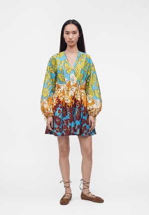 Žena s dlhými čiernymi vlasmi stojaca, oblečená v farebných kvetinových šatách s nazdobenými rukávmi a hnedých viazaných balerínach na bielom pozadí.