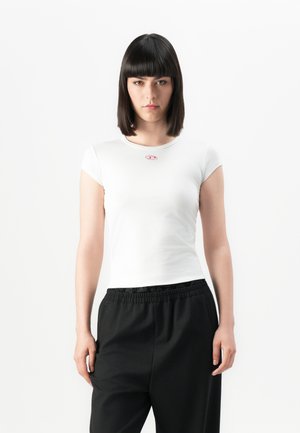 Woman with straight dark hair wearing a fitted white short-sleeve top and loose black pants, standing against a plain white background.