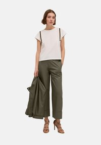 Top alb cu mâneci scurte și accente negre, asortat cu pantaloni lungi de culoare verde măsliniu. Modelul ține o jachetă asortată de culoare verde măsliniu.