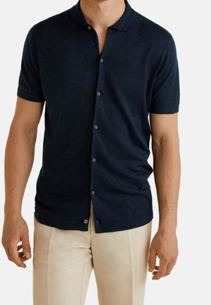 Hombre con camiseta tipo polo de manga corta azul marino y pantalones beige claros, de pie frente a un fondo liso.