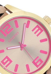 Montre ronde en or avec un cadran argenté, des index heures roses et des aiguilles. Bracelet texturé avec un imprimé animal. Arbore le logo de la marque "oozoo."