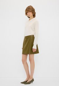 Blusa bianca a maniche lunghe con colletto abbinata a una gonna a trapezio in similpelle verde con dettagli laterali, indossata con scarpe verdi con tacco basso.