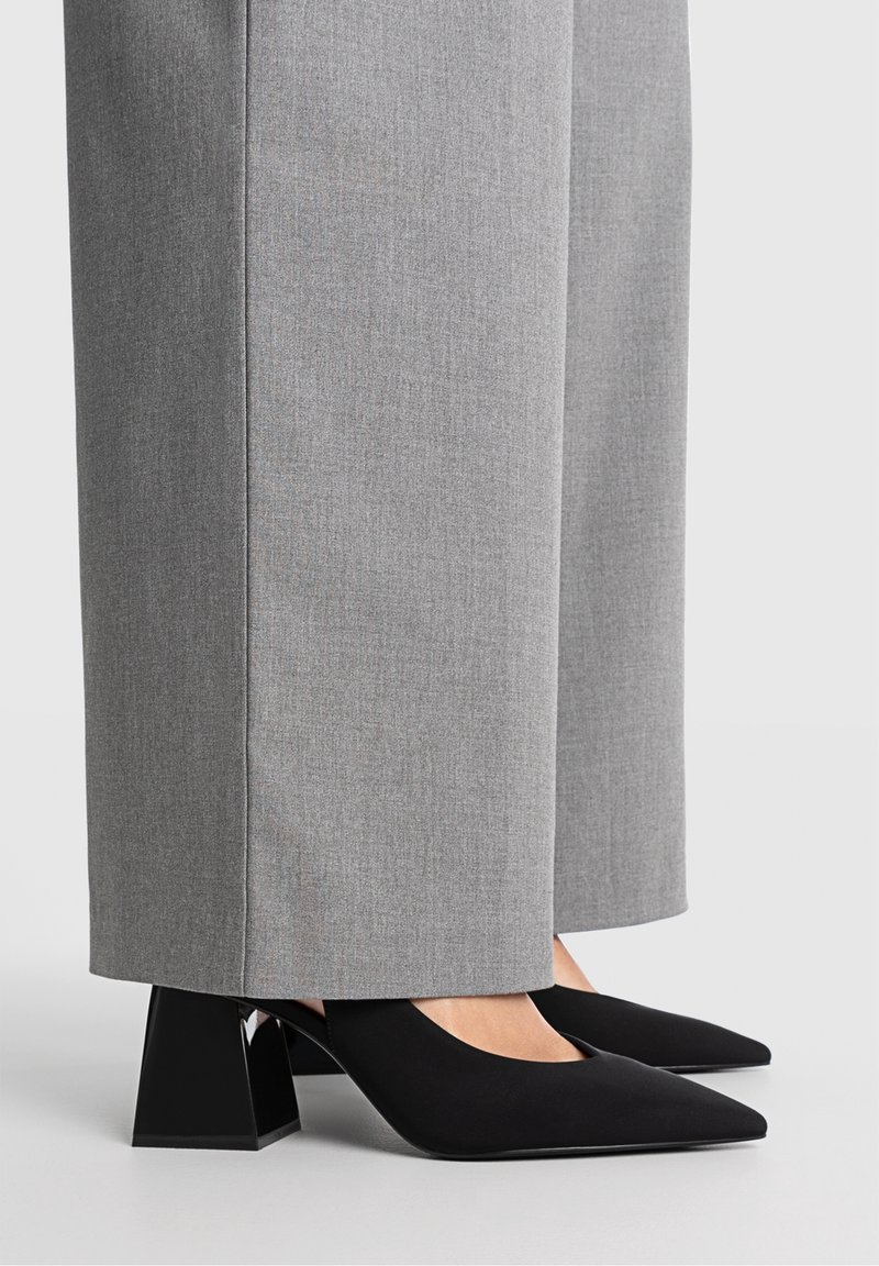 Personne portant des chaussures noires à talons carrés pointus associées à un pantalon gris à jambes larges, debout sur une surface grise claire unie.