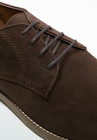 Chaussure en suède marron avec un bout arrondi, des lacets gris et des coutures détaillées sur le dessus. Présente une semelle de couleur claire.