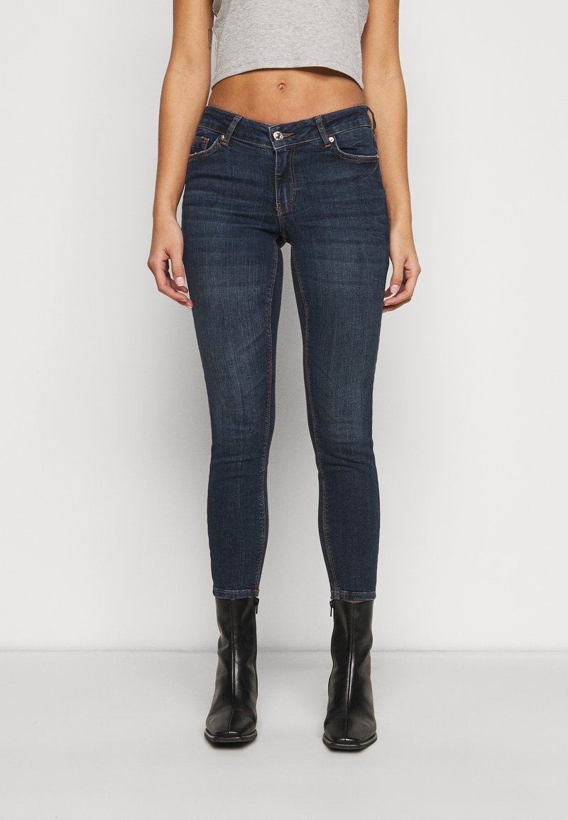Jeans skinny azul escuro com uma textura suave e costuras contrastantes, combinados com botas pretas até o tornozelo. Apresenta um fecho de botão e zíper.