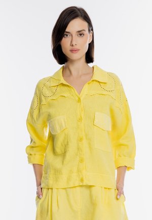 Kobieta z krótkimi ciemnymi włosami, ubrana w żółtą koszulę zapinaną na guziki z kieszeniami i ozdobnymi oczkami, stoi z rękami w kieszeniach na białym tle.