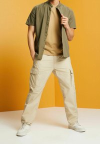 Man in beige cargobroek, lichtbruine T-shirt en olijfgroen kortebloes met knopen, staande tegen een oranje achtergrond.