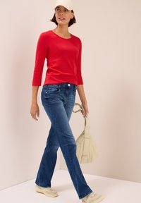 Rotes, figurnahes Langarmshirt kombiniert mit blauen Schlagjeans. Das Model trägt eine hellbeige Tasche, kremfarbene Sneakers und eine weiße Mütze.