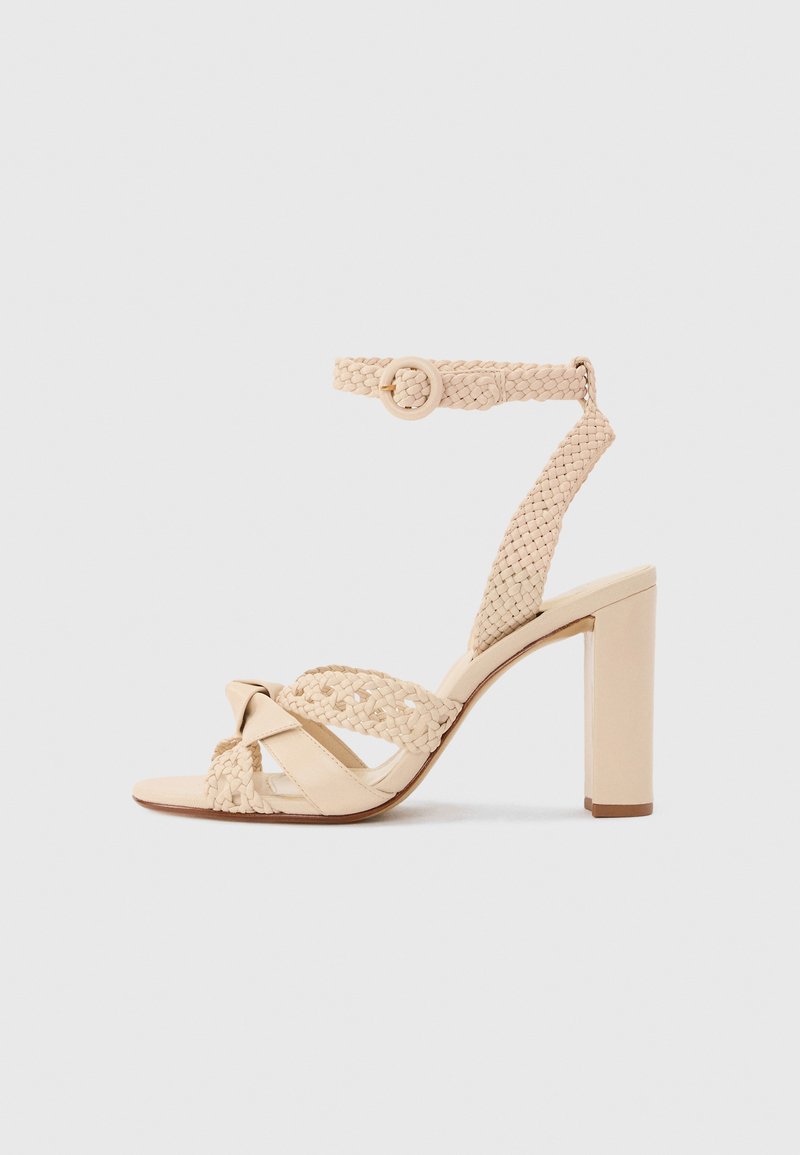 Μπεζ ψηλοτάκουνο σανδάλι με πλεγμένα λουριά, πόρπη στον αστράγαλο, ανοιχτή μύτη και χοντρό τακούνι σε απλό ανοιχτό γκρι φόντο.