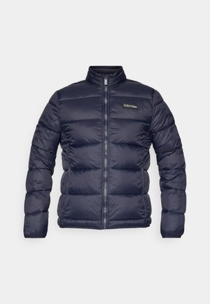 Marinblå puffjacka med dragkedja, horisontellt kviltade paneler och ståkrage. Har en liten logopatch på vänster bröst.