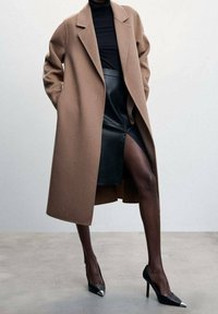 Manteau en laine marron avec une coupe décontractée, associé à une jupe en cuir noir et des talons aiguilles noirs avec des embouts métalliques. Texture lisse et lignes épurées.