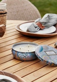 Doftljus i en rund, dekorativ burk med blå och rosa blommönster. Tre vekar är synliga. Placerad på ett träbord.