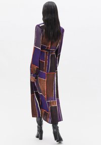 Abito lungo viola con un motivo geometrico arancione e marrone, maniche lunghe e vita cinchata, abbinato a stivali neri al ginocchio.