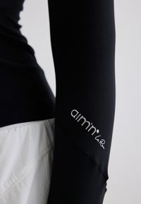 Gros plan d'une personne portant un haut à manches longues noir avec le logo "aimn" sur la manche et un pantalon blanc texturé.