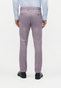 Moški v ozkih vijoličastih hlačah za obleko, svetlo modri srajci in črnih elegantnih čevljih, stoji z obrazi zadaj proti gledalcu.