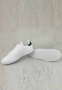 Sneakers bianchi con superficie testurizzata, accentuazione verde nel tallone e suola bianca con motivo esagonale. Presentano lacci piatti e un design a profilo basso.