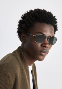 Sunglasses featuring a silver metal frame, aviator shape, and dark tinted lenses. Styled with a ribbed brown bomber jacket.