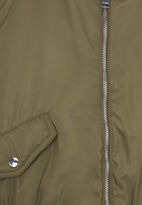 Acercamiento de una chaqueta verde oliva con cremallera plateada y un bolsillo con tapa abotonada en el lado izquierdo.