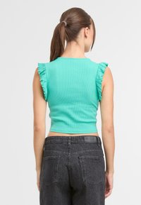 Kobieta z brązowym kucykiem, ubrana w bezrękawnik w turkusowym kolorze z falbankami na ramionach oraz czarne dżinsy z wysokim stanem, zwrócona plecami.