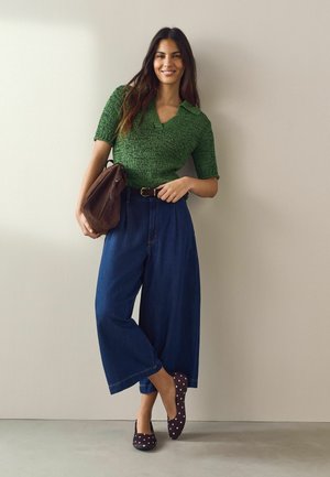 Woman with long dark hair wearing a green knit top, wide blue cropped jeans, and maroon polka dot flats, holding a brown leather bag.