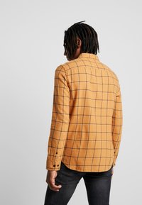 Camicia a maniche lunghe in tessuto giallo dorato con motivo a quadri in marrone scuro. Presenta polsini con bottoni e colletto a punta.