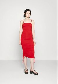 Jovem mulher de pé, vestindo um vestido midi vermelho ajustado com alças finas e sandálias de plataforma pretas, contra um fundo claro e simples.