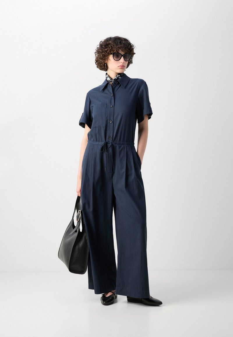 Navyblauer Jumpsuit mit kurzen Ärmeln, Knopfverschluss und Kordelzug in der Taille; kombiniert mit einer schwarzen Handtasche und spitzen Schuhen.