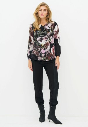 Woman wearing floral blouse with long sleeves, black fitted pants, and black heeled ankle boots standing against white background.