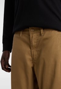 Brązowe bawełniane spodnie o prostym kroju, z pojedynczym zapięciem na guzik, kieszeniami bocznymi i gładkim wykończeniem. Noszone z czarną koszulą z długim rękawem.