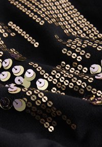 Črna tkanina, okrašena z vzporedno prekrivajočimi se metalnimi zlatimi in srebrnimi bleščicami, ki so razporejene v vzorec in ustvarjajo teksturiran bleščeči učinek.