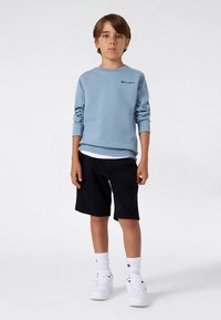 Zēns stāv taisni, ģērbies gaiši zilā džemperī, melnās šortos, balto augsto zeķēs un baltās kedās uz vienkārša fona.