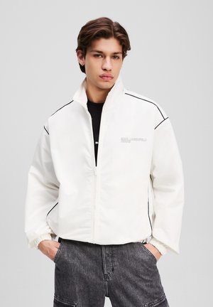 White zip-up jacket with black side stripes and a high collar. Features elastic cuffs and a subtle logo on the front. Smooth texture.