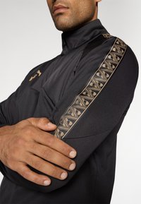 Chaqueta atlética negra con cuello alto, material texturizado en negro y una franja decorativa con un patrón dorado a lo largo del hombro.