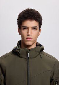 Jaunas vyras su trumpais garbanotais plaukais, dėvintis užtrauktą alyvuogių žalios spalvos gaubtuotą striukę, priešais paprastą šviesią foną.