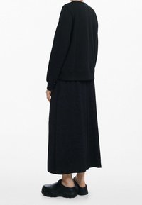 Sudadera de algodón negra con cuello redondo, mangas largas y dobladillo elástico, combinada con una falda negra hasta el tobillo y zuecos negros de suela gruesa.