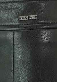 Čierny textúrovaný kožený materiál s logom GUESS na doske, so viditeľným šitím a hladkým povrchom.
