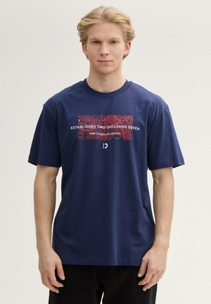 T-shirt en coton bleu marine avec un design graphique rouge et du texte, à manches courtes et encolure ronde. Porté avec un pantalon noir.